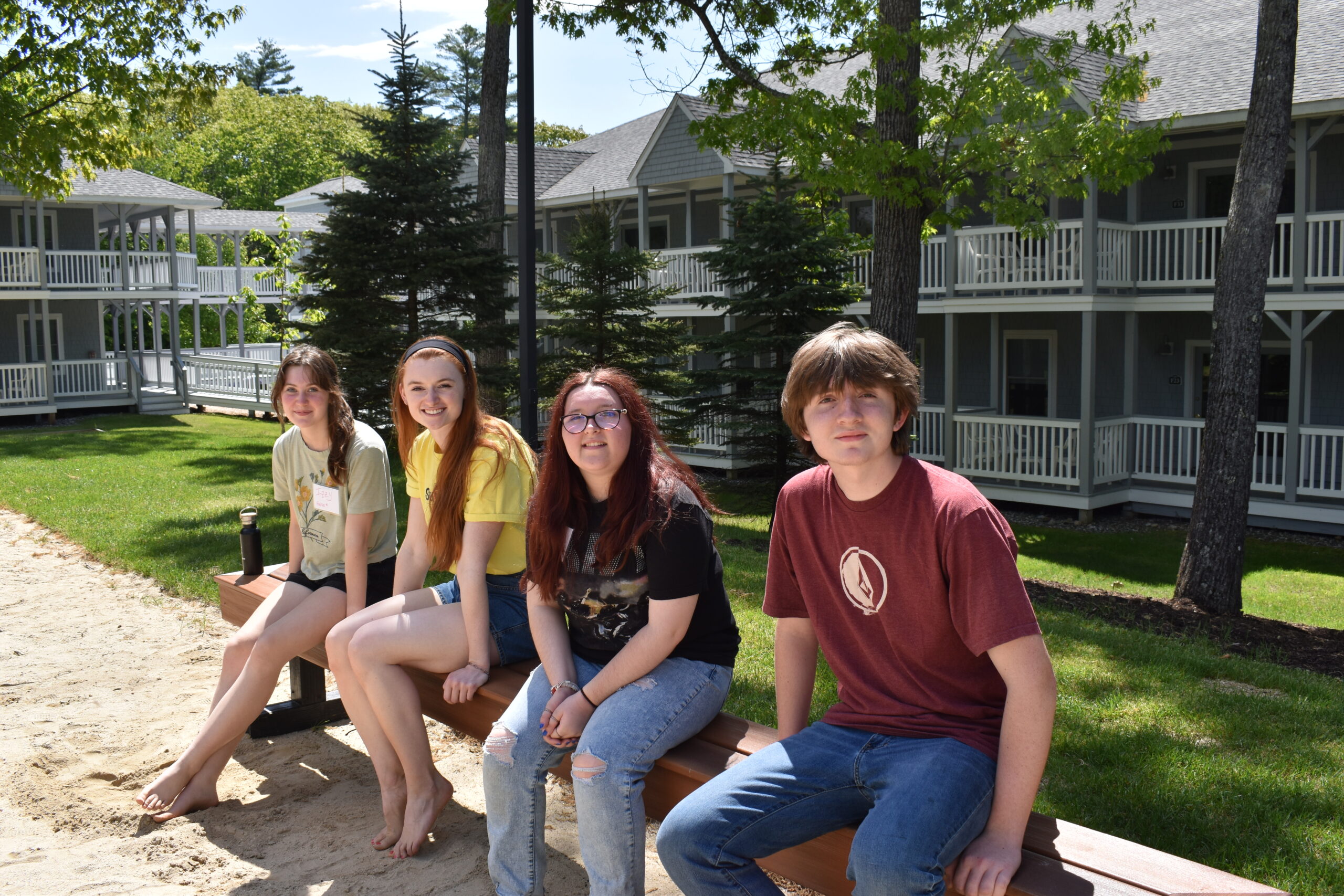 a group of four young adults sit on a bench on the side of a beach volleyball court. There are many rows of camp cabins behind them. All of the young adults are white, and one girl has brown hair, two have red hair and one boy has medium length brown hair.